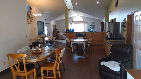Open common area with multiple dining tables and chairs, a recliner, and a reception counter under a vaulted ceiling and arched window.