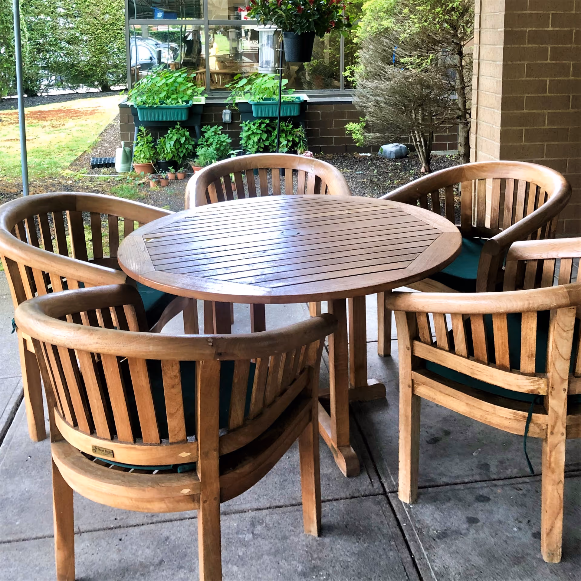 Outdoor patio area with a round wooden table surrounded by five wooden chairs with green cushions. In the background, there are green plants in pots and planters against a brick wall and a window reflecting the outdoor scene.