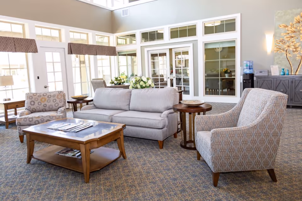 Bright furnished common lounge with a sofa, patterned armchairs, coffee table, and large windows.