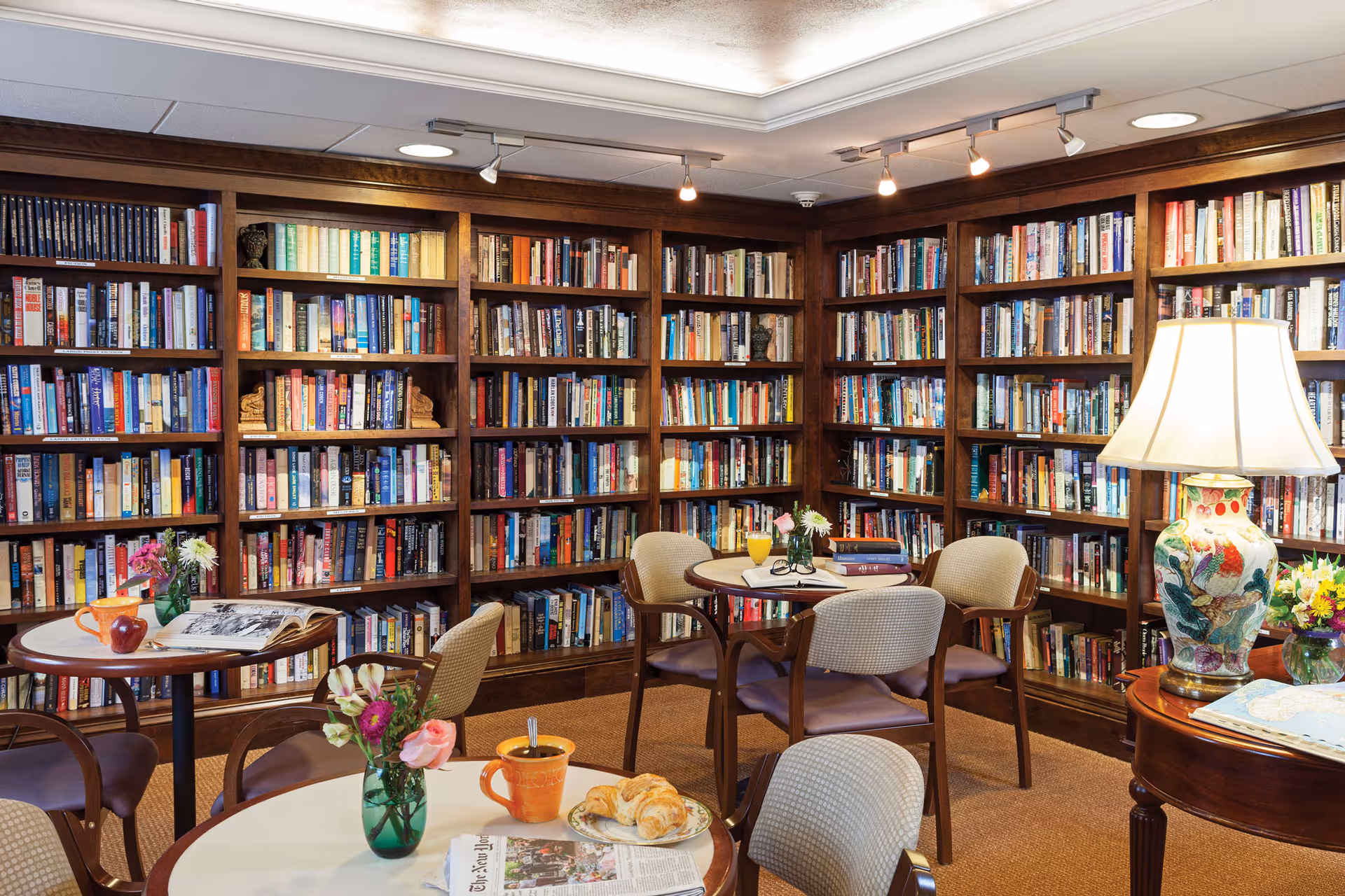 Cozy library room with wooden bookshelves filled with books lining the walls. Several round tables with chairs are arranged in the room, each table decorated with small flower vases, books, and refreshments including a croissant, coffee mugs, and a glass of orange juice. A decorative table lamp with a floral design is placed on a side table.