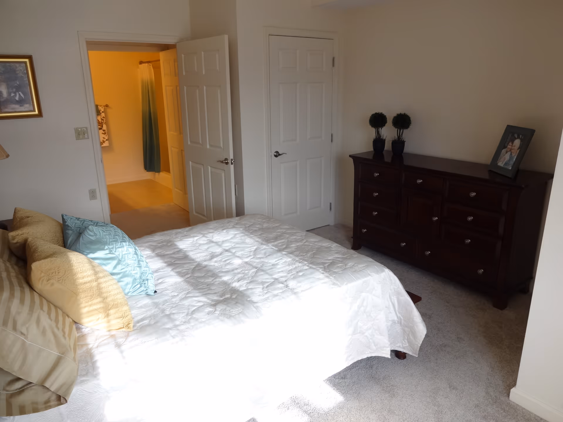 Sunlit bedroom with a bed and pillows, a dark wood dresser, and an open door leading to a bathroom.