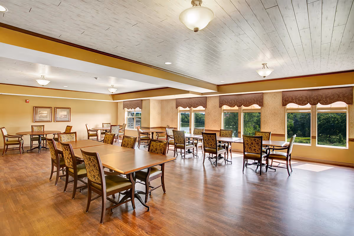 Spacious dining room with multiple wooden tables and upholstered chairs, large windows overlooking greenery, and warm overhead lighting.