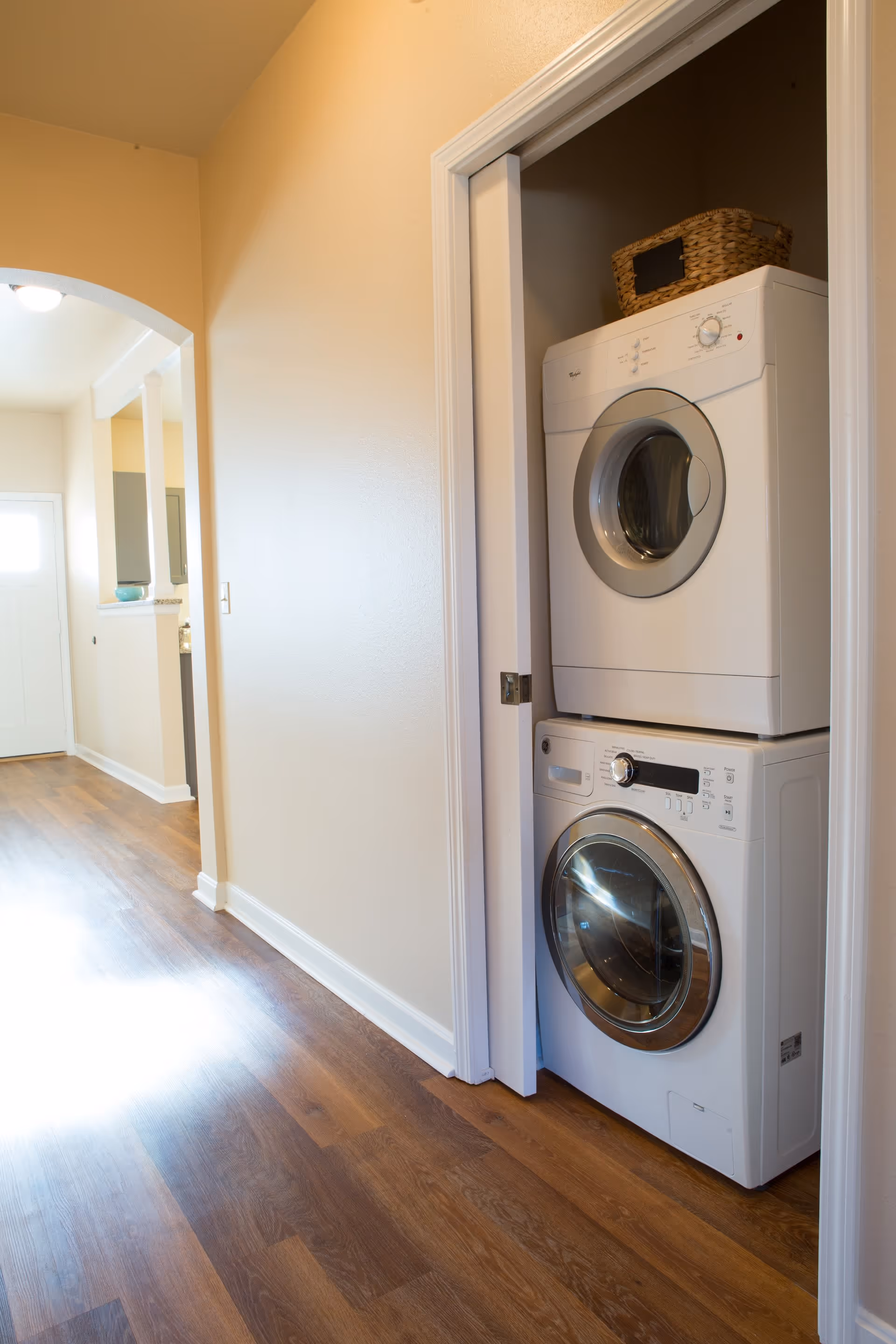 A hallway with wooden flooring leading to a white door, featuring a stacked washer and dryer unit inside a small closet with a woven basket on top.