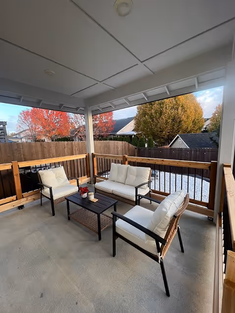 Covered outdoor patio area with cushioned seating including a loveseat and two armchairs arranged around a small black coffee table. The patio is enclosed with wooden railings and overlooks a fenced backyard with trees showing autumn foliage.