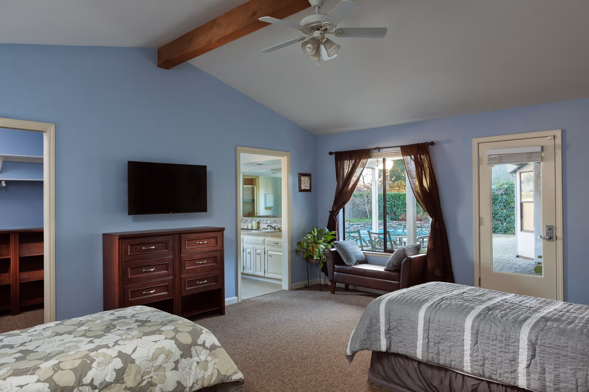 Light blue-walled bedroom with two beds, a wall-mounted TV over a wooden dresser, a small seating area by a window, and doors to an en-suite bathroom and patio.