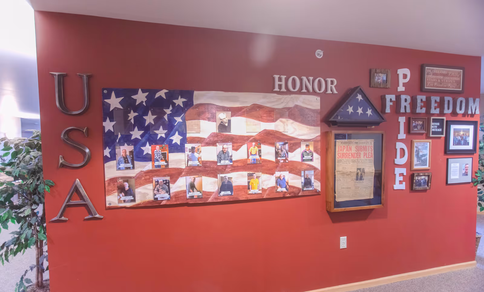 A red wall display honoring veterans with a large painted American flag, photos of veterans, and words such as USA, HONOR, PRIDE, and FREEDOM. There is a folded flag in a triangular wooden case and a framed newspaper clipping titled 'Japan Submits Surrender Plea'. Various framed photos and patriotic decorations are also visible.