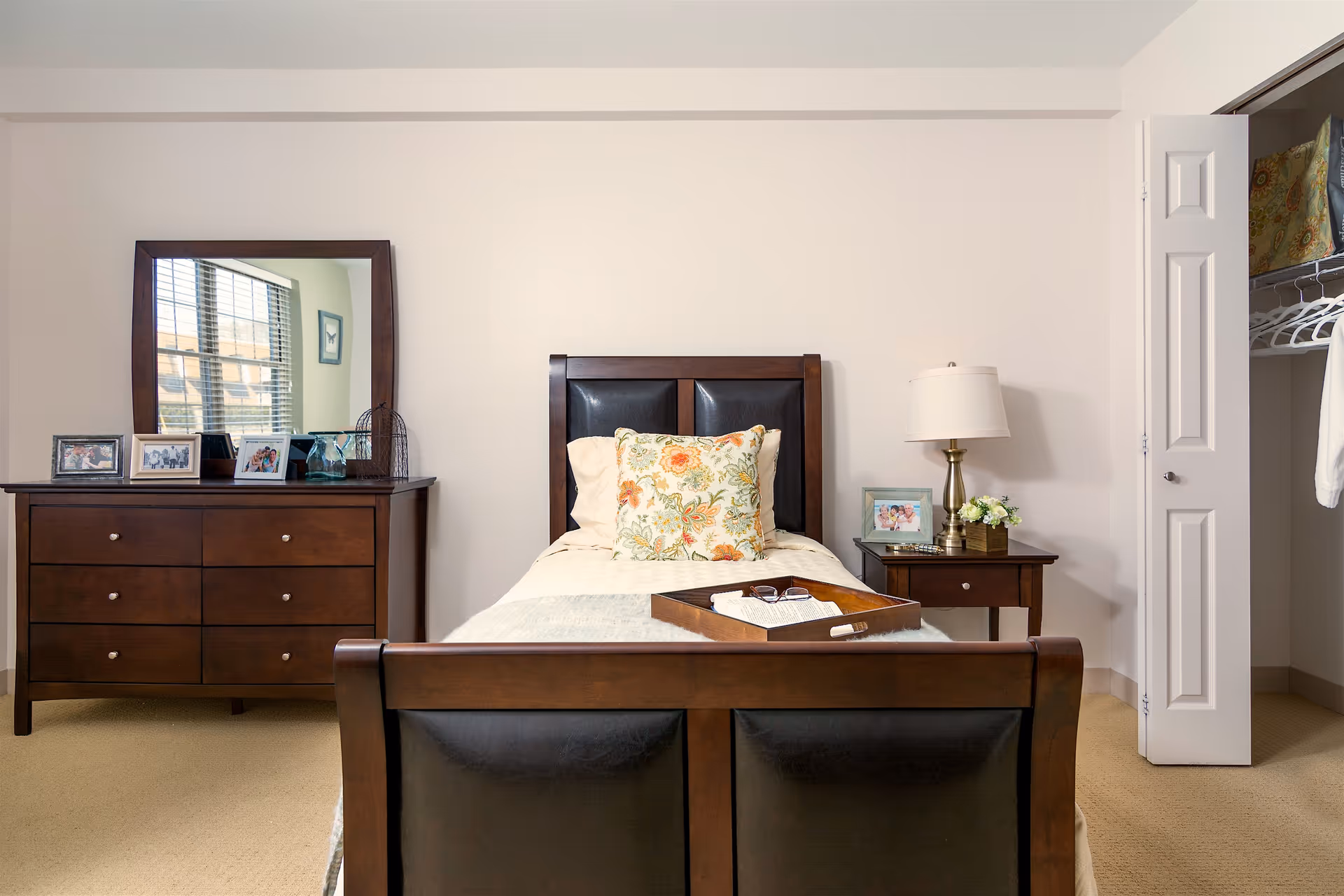A neatly arranged bedroom with a single wooden bed, dresser with mirror, bedside table and lamp, and an open closet.