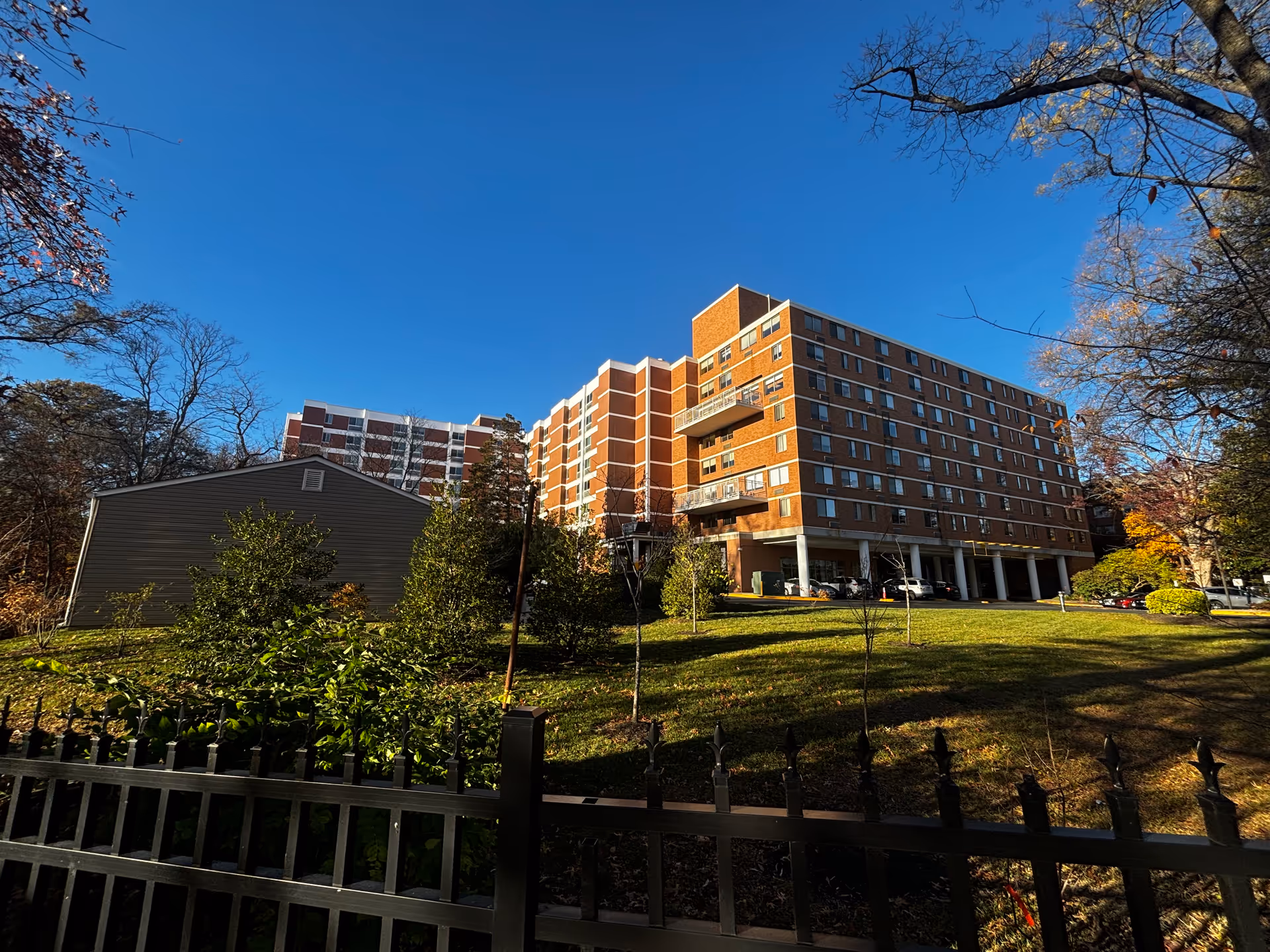 Exterior view of a multi-story brick residential building with balconies, surrounded by trees and greenery under a clear blue sky. A black metal fence is visible in the foreground.