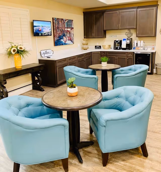 A cozy seating area with two round tables, each surrounded by two light blue tufted armchairs. The background features a kitchenette with dark wood cabinets, a coffee machine, and a small refrigerator. There is a decorative painting on the wall and a small table with a vase of sunflowers near a window with white shutters.