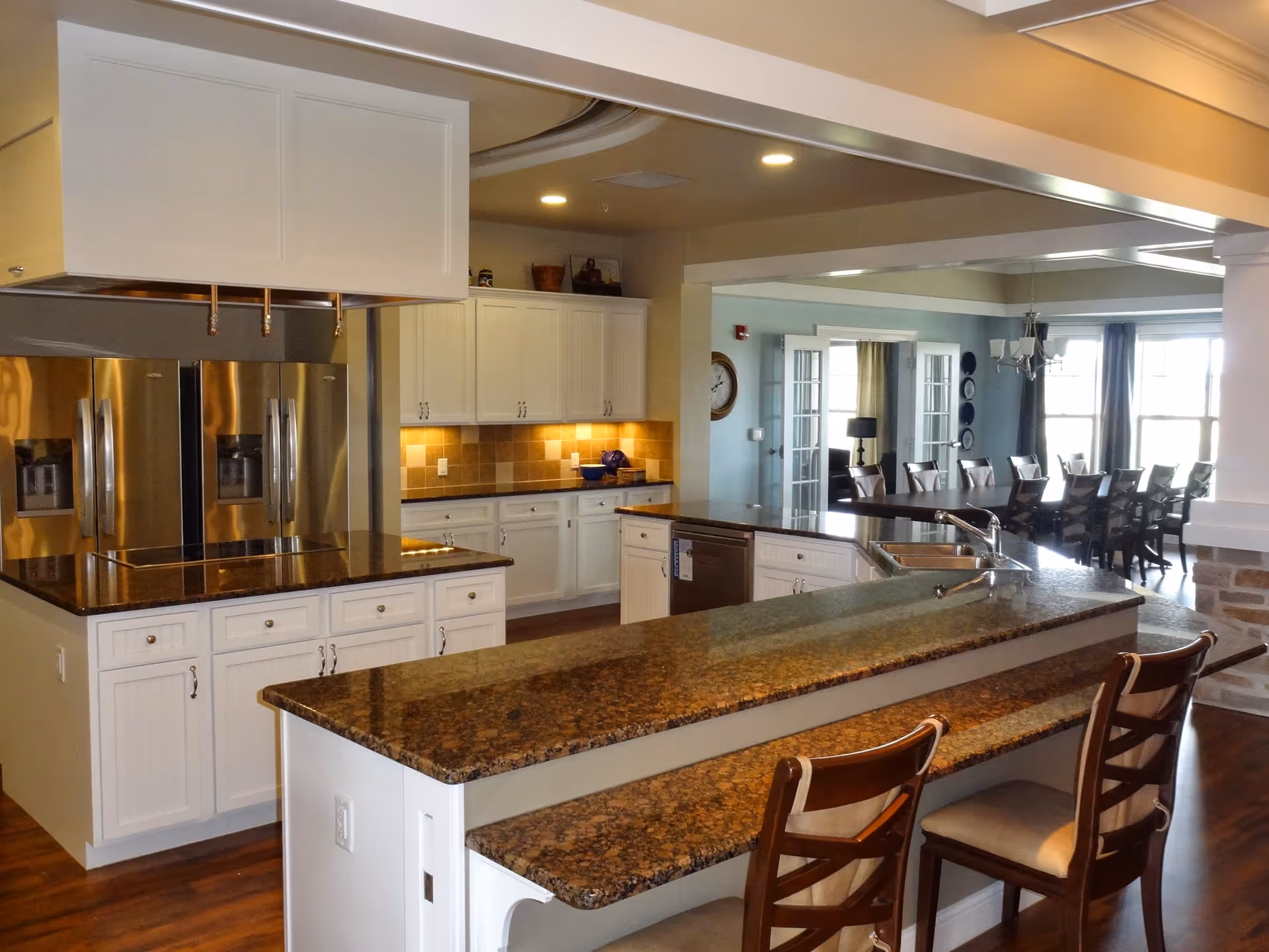 A spacious kitchen area with granite countertops, white cabinetry, and stainless steel appliances including a double-door refrigerator. There is a kitchen island with a sink and two wooden chairs with cushioned seats. In the background, a dining area with a long table and multiple chairs is visible, illuminated by natural light from large windows.
