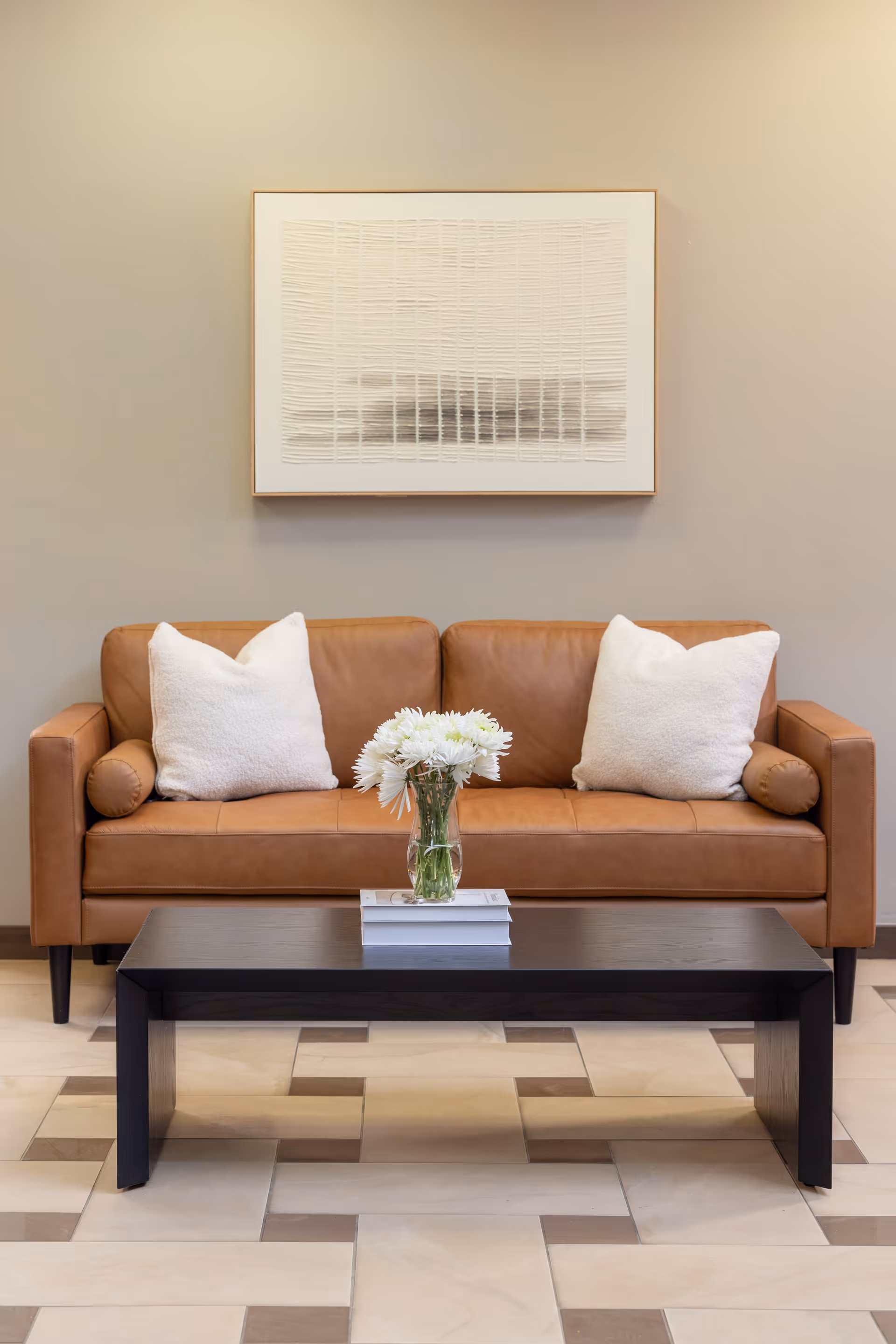 A tan leather sofa with white pillows and a black coffee table holding a vase of white flowers beneath framed wall art.
