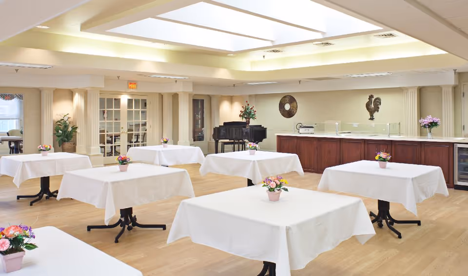Bright dining/activity room with multiple tables draped in white cloths and small floral centerpieces, a piano and serving counter in the background.