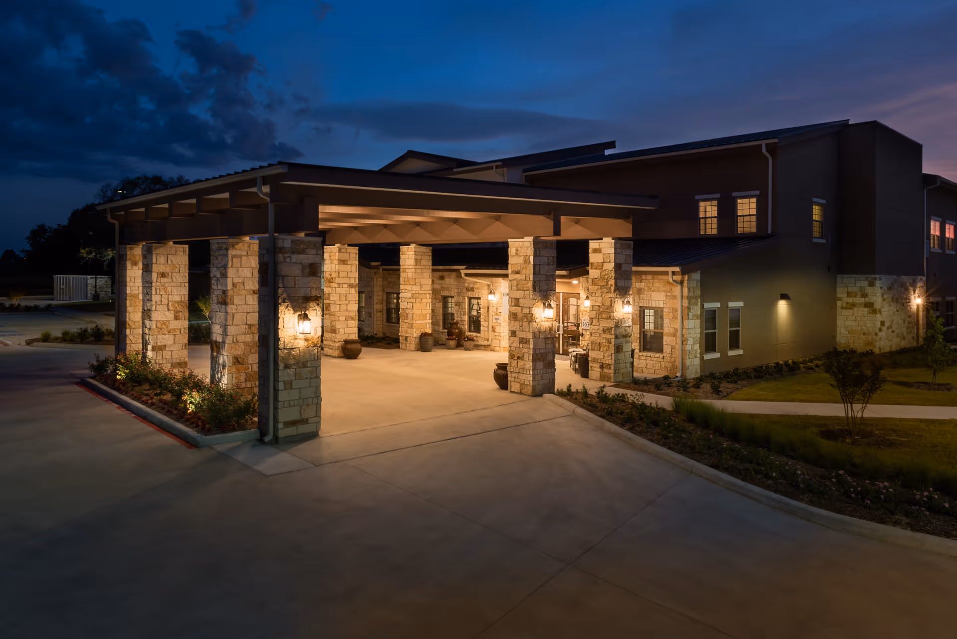 Covered entrance with stone columns and warm exterior lights at the front of a senior living building at dusk.