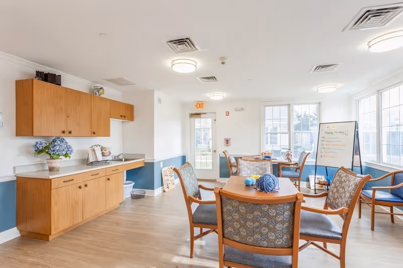 Bright communal activity/dining room with a kitchenette on the left, tables and chairs in the center, and large windows letting in natural light.