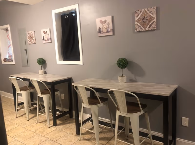 Interior view of a room with two narrow tables against a gray wall, each table accompanied by two metal chairs with wooden seats. Small potted topiary plants are placed on each table. The wall above the tables is decorated with three small framed artworks. The floor is tiled in a light beige color.