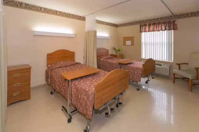 A healthcare facility room with two hospital beds separated by a privacy curtain. Each bed has a brown patterned bedspread and an overbed table. There is a wooden nightstand with drawers next to one bed, a window with vertical blinds and a valance, an air conditioning unit below the window, a corkboard on the wall, and a beige armchair in the corner.