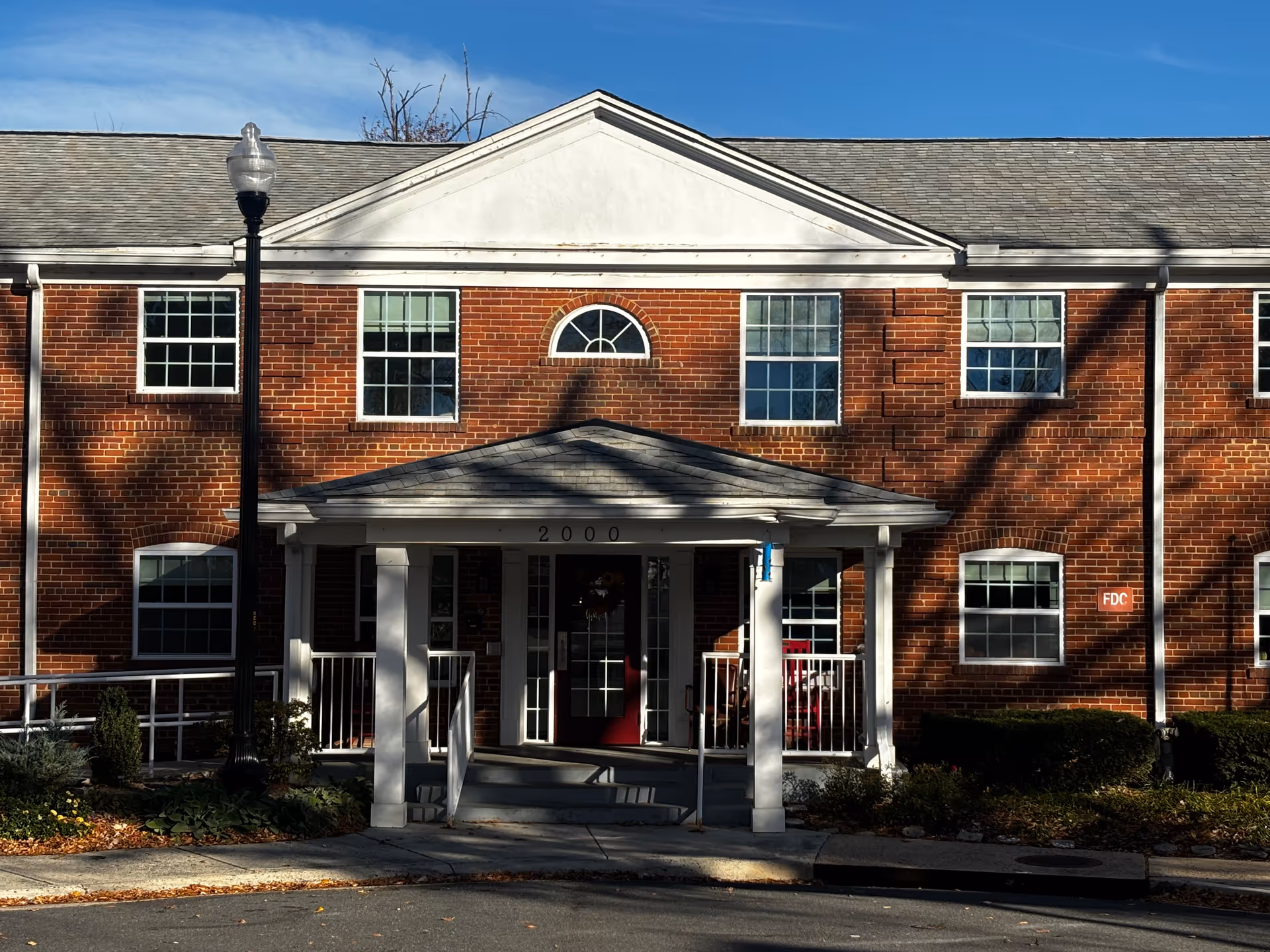 Front exterior view of a two-story brick building with white trim and a small covered entrance porch. The building has multiple windows and a street lamp in front. The number 2000 is displayed above the entrance door.