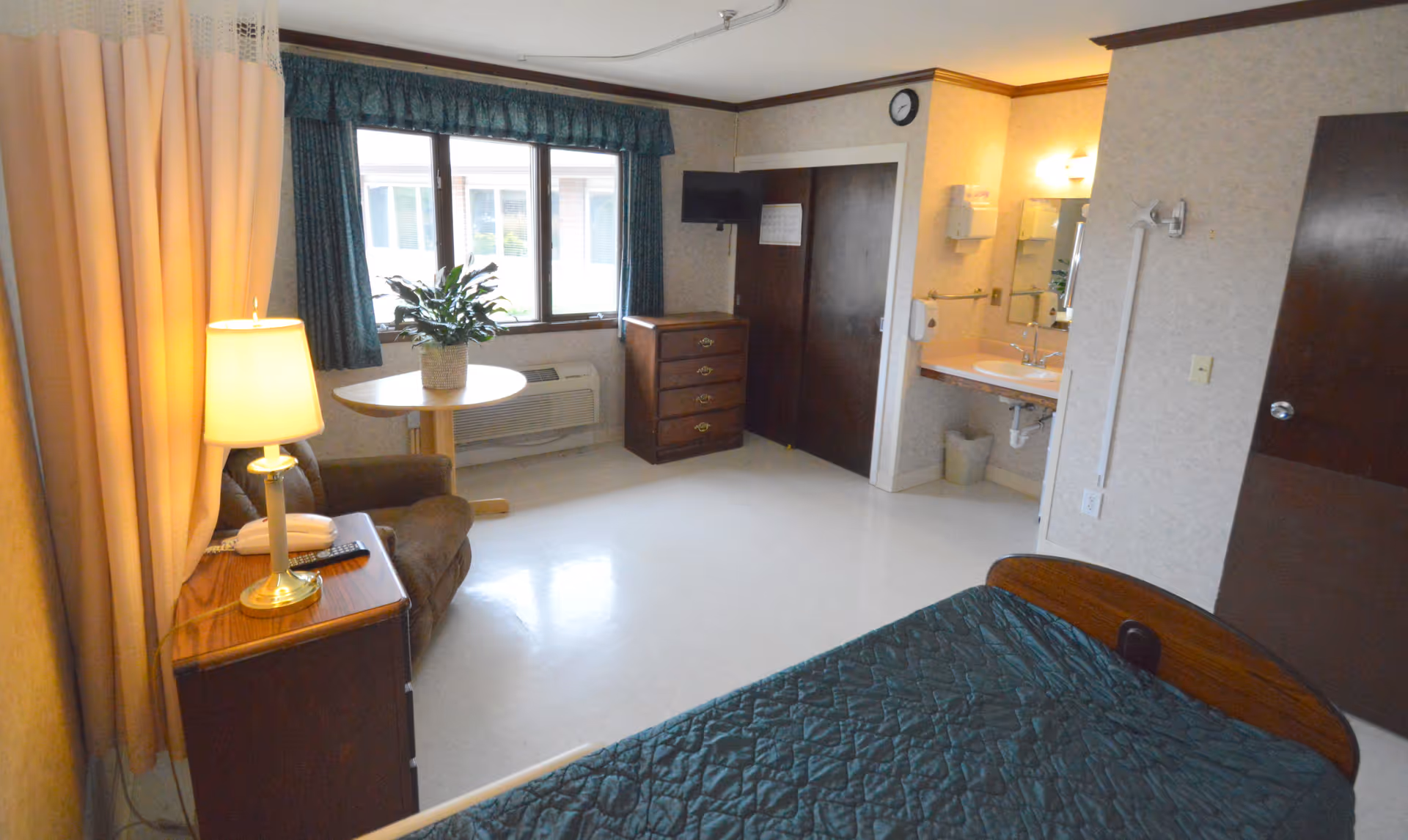 Private resident bedroom in a senior care facility featuring a bed, armchair, dresser, small table by a window and an open sink area.