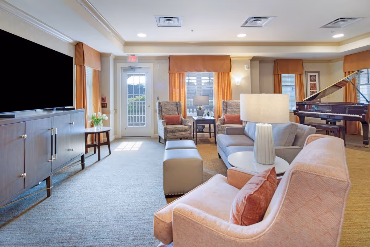 Bright, well-furnished living room with sofas, armchairs, a piano, large TV, and windows with orange curtains.