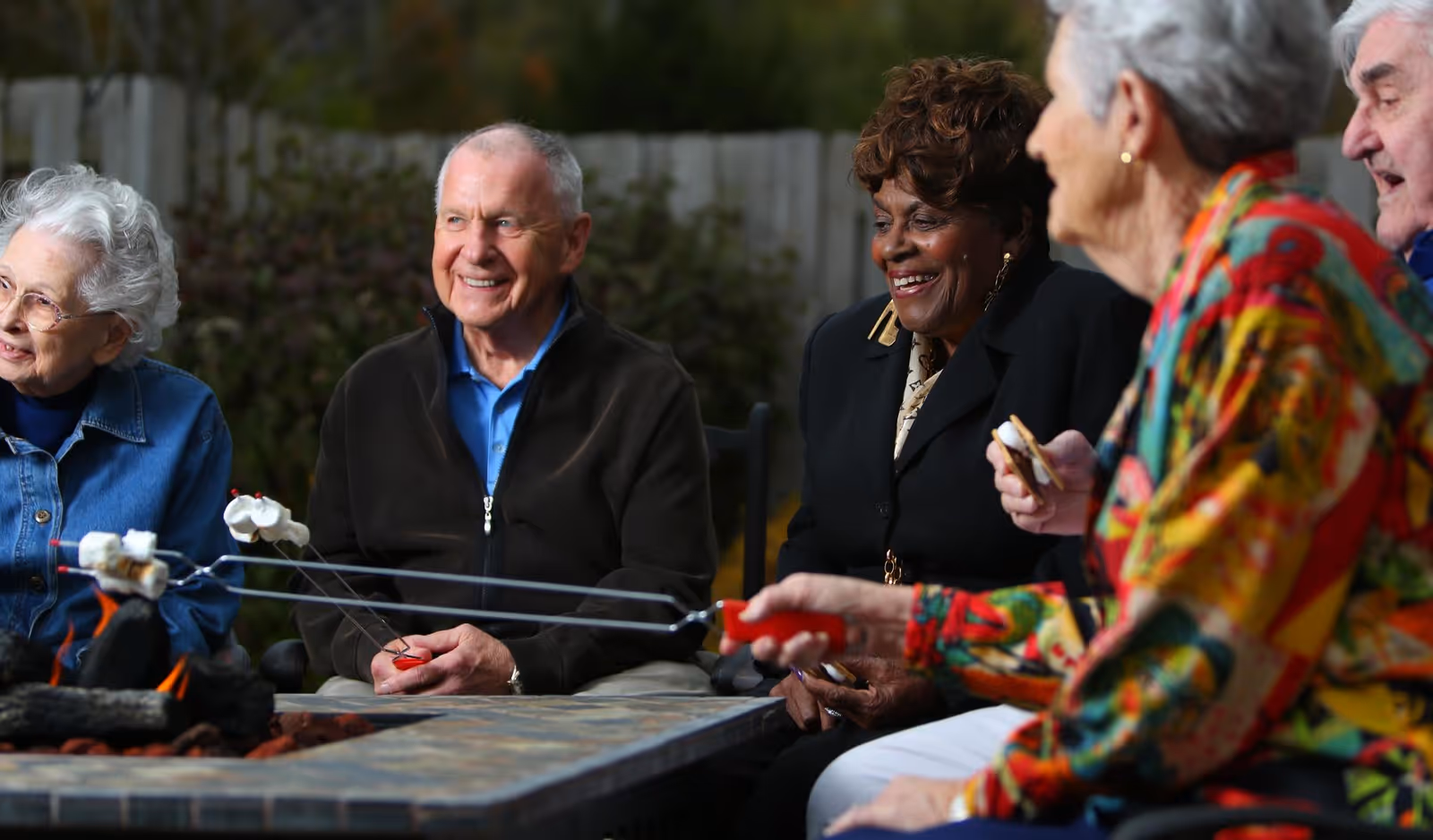 A group of elderly people sitting outdoors around a fire pit, roasting marshmallows and smiling, enjoying each other's company.