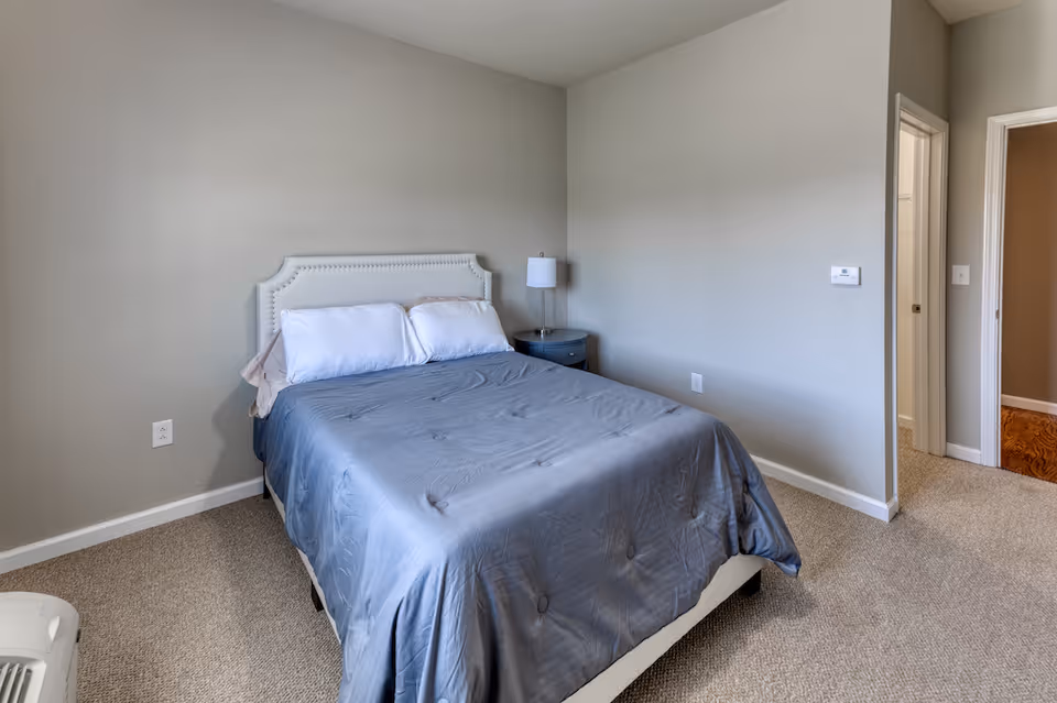 A simple bedroom with a bed covered in a gray comforter and two white pillows. There is a small round bedside table with a lamp on it next to the bed. The walls are painted light gray and the floor is carpeted. Two doorways are visible, one leading to another room with wooden flooring.