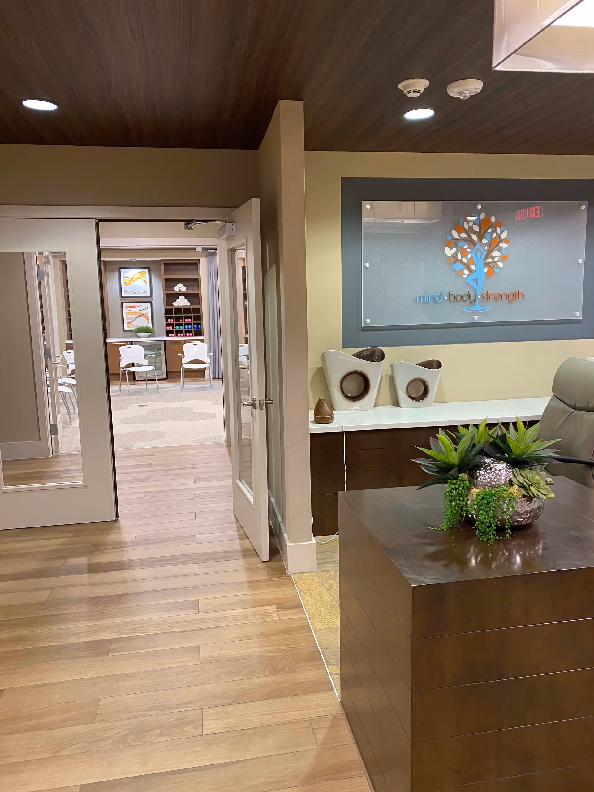 Interior view of a senior living facility showing a wooden floor and a reception desk with a plant arrangement. In the background, there is an open door leading to a room with chairs and shelves holding towels and exercise equipment. A wall sign reads 'mind body strength' with a tree logo.