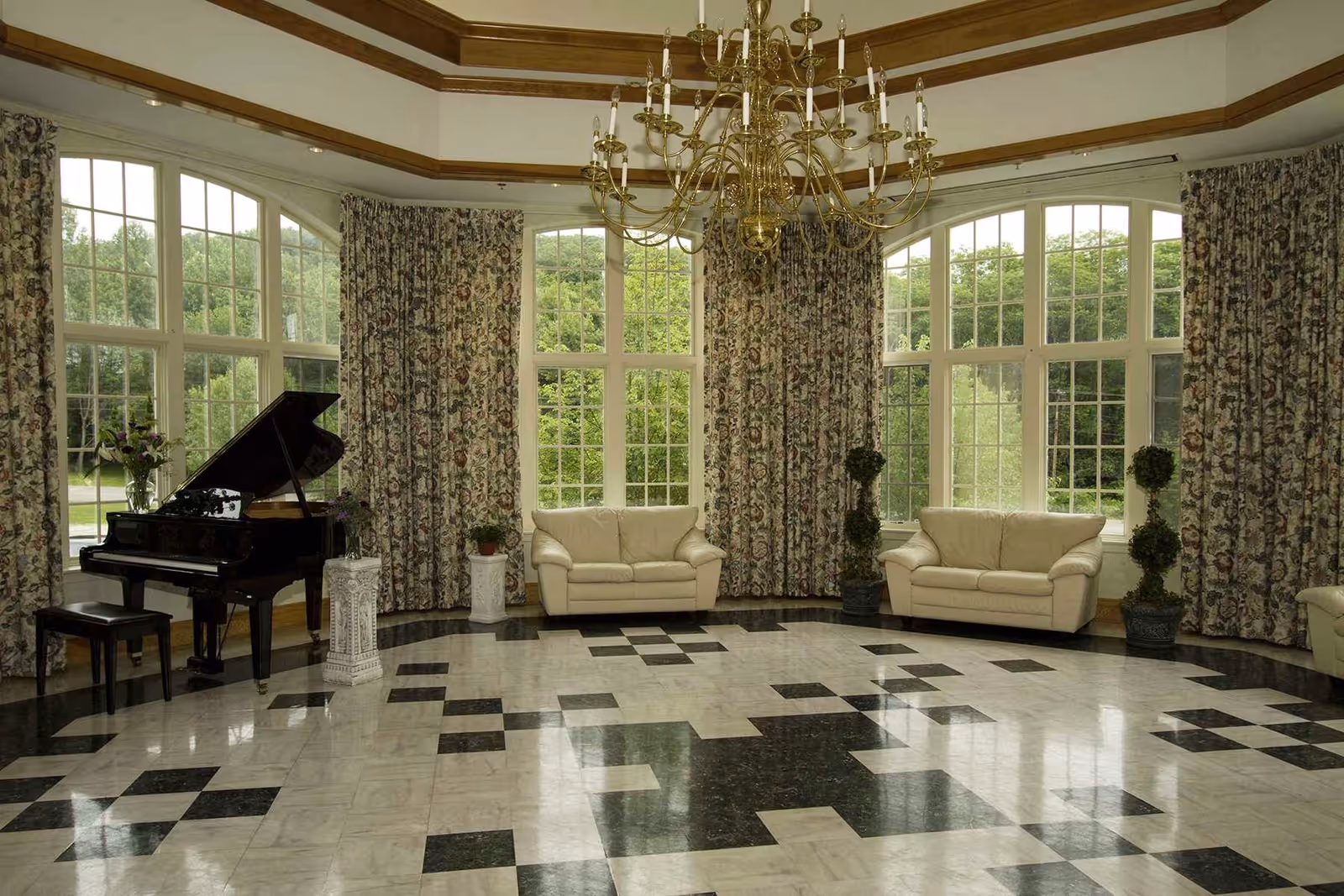 A spacious living room with large windows covered by floral curtains, two cream-colored sofas, a black grand piano with a matching bench, decorative plants, and a large gold chandelier hanging from the ceiling. The floor features a black and white checkered pattern.