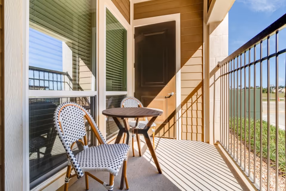 Sunlit balcony with two woven chairs and a small round table next to a door and metal railing.
