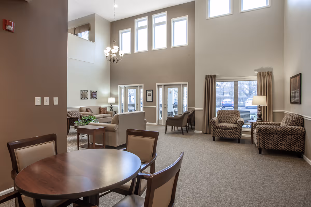 A spacious living room area in a senior living facility with high ceilings and multiple windows letting in natural light. The room features a round wooden table with four chairs in the foreground, several upholstered armchairs and sofas arranged around side tables and lamps, beige carpeting, and neutral-colored walls. There are framed pictures on the walls and a chandelier hanging from the ceiling.