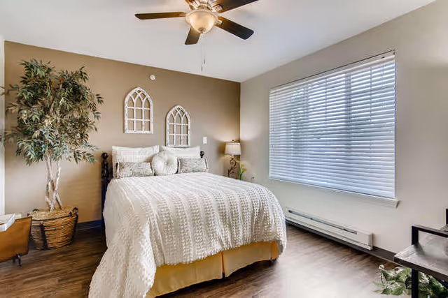 A cozy bedroom with a neatly made bed featuring a textured white comforter and multiple pillows. The room has a large window with closed blinds, a ceiling fan with a light, a potted plant in a woven basket, and two decorative arched wall hangings above the bed. The floor is wooden, and there is a small table with a lamp and a plant beside the bed.