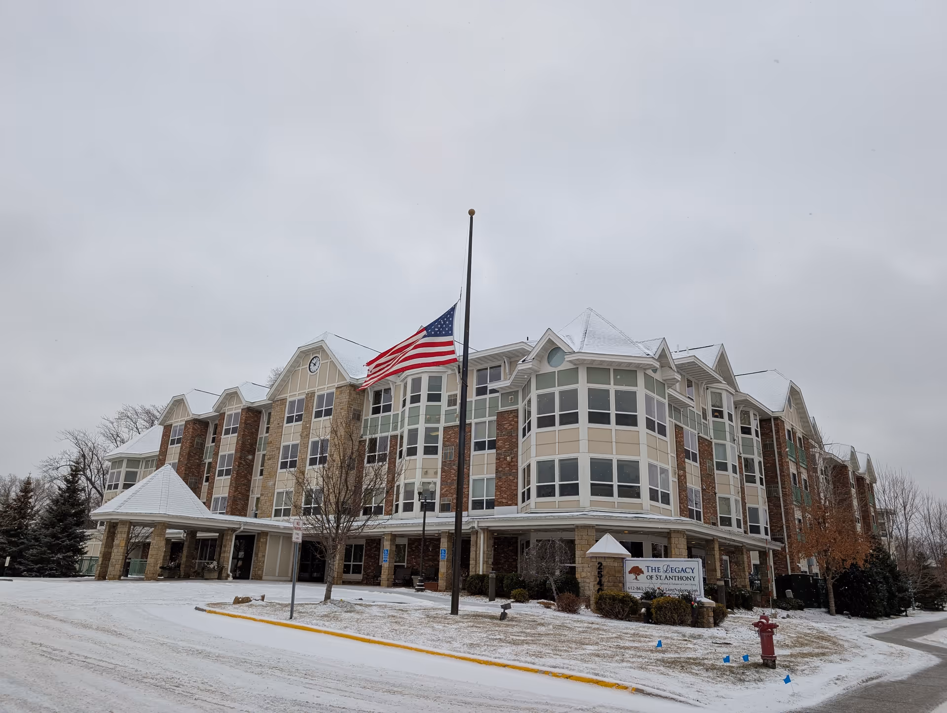 Front exterior of a multi-story senior living building with an American flag at half-mast and light snow on the ground.