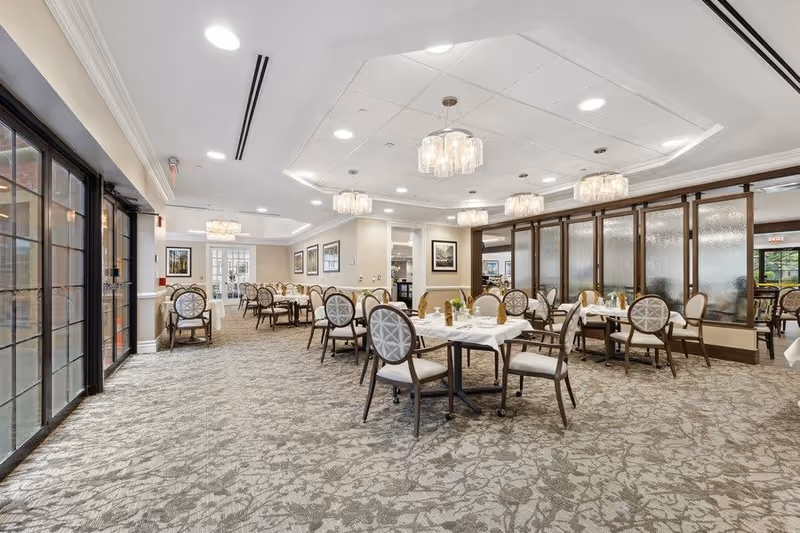 A spacious and elegant dining room with multiple round tables covered with white tablecloths, each set with napkins and glassware. The room features patterned carpet flooring, large windows on the left side, framed artwork on the walls, and several modern chandeliers hanging from the ceiling. There are cushioned chairs around each table, and a partition with frosted glass panels separates part of the room.