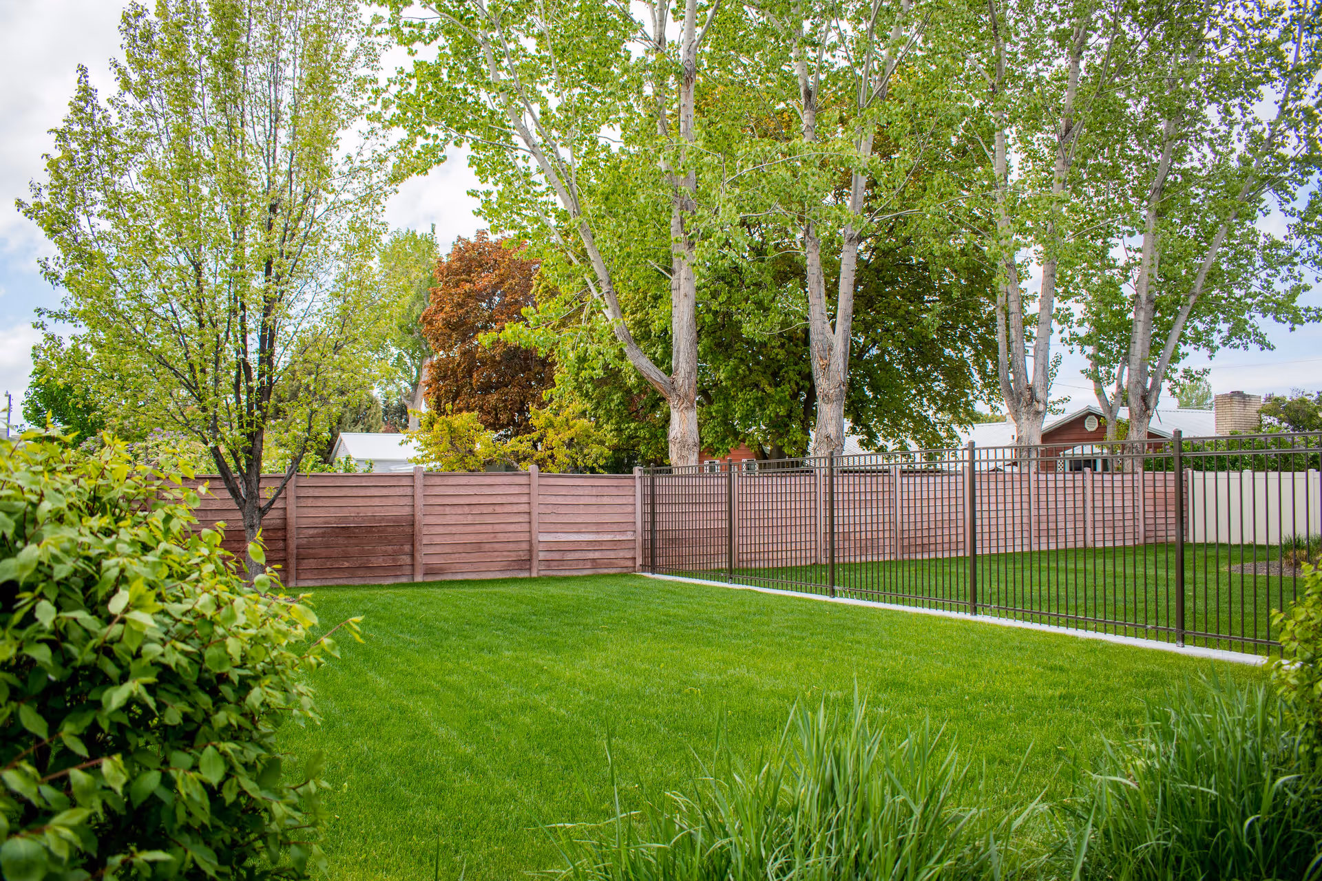 A well-kept grassy yard with a wooden and metal fence, trees, and shrubs.