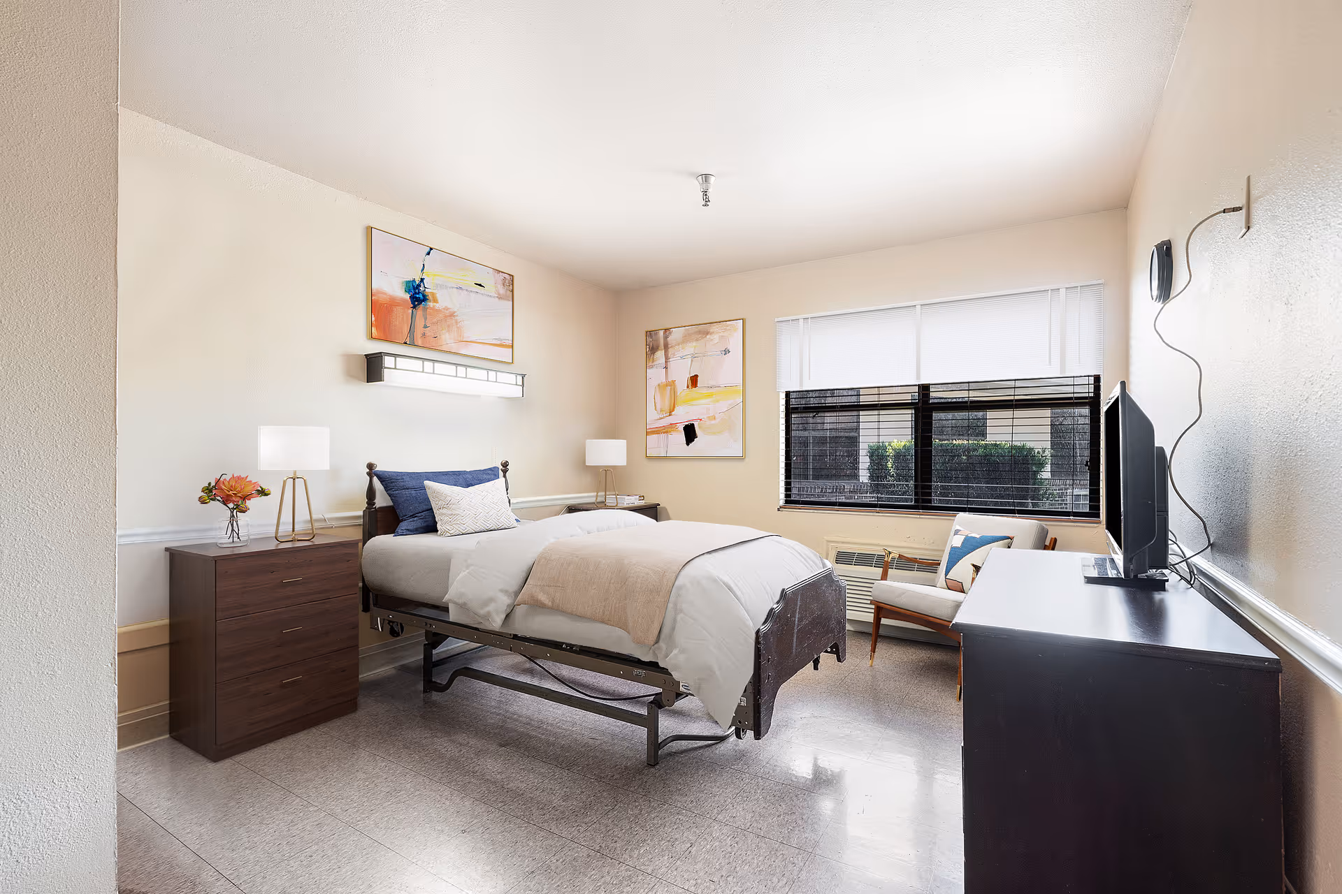 Bright, neatly arranged single-occupancy bedroom with a hospital-style bed, dresser, TV, armchair, and large window.