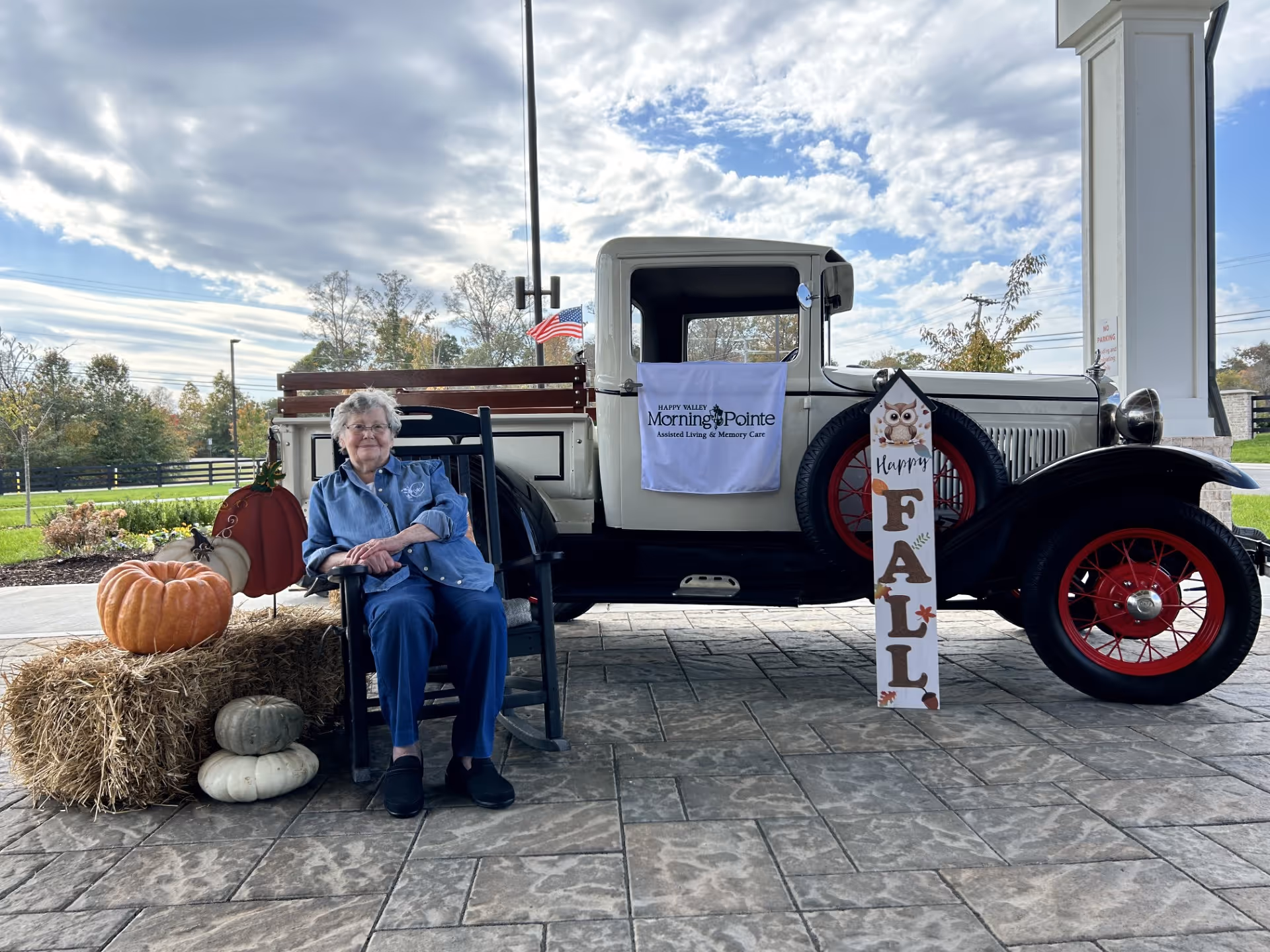 An elderly woman sitting in a black rocking chair next to a vintage white truck with red wheels. The truck has a sign on its door that reads 'Happy Valley Morning Pointe Assisted Living & Memory Care.' There are fall decorations including pumpkins, hay bales, and a vertical sign with an owl that says 'Happy FALL.' The scene is outdoors with a partly cloudy sky and some greenery in the background.