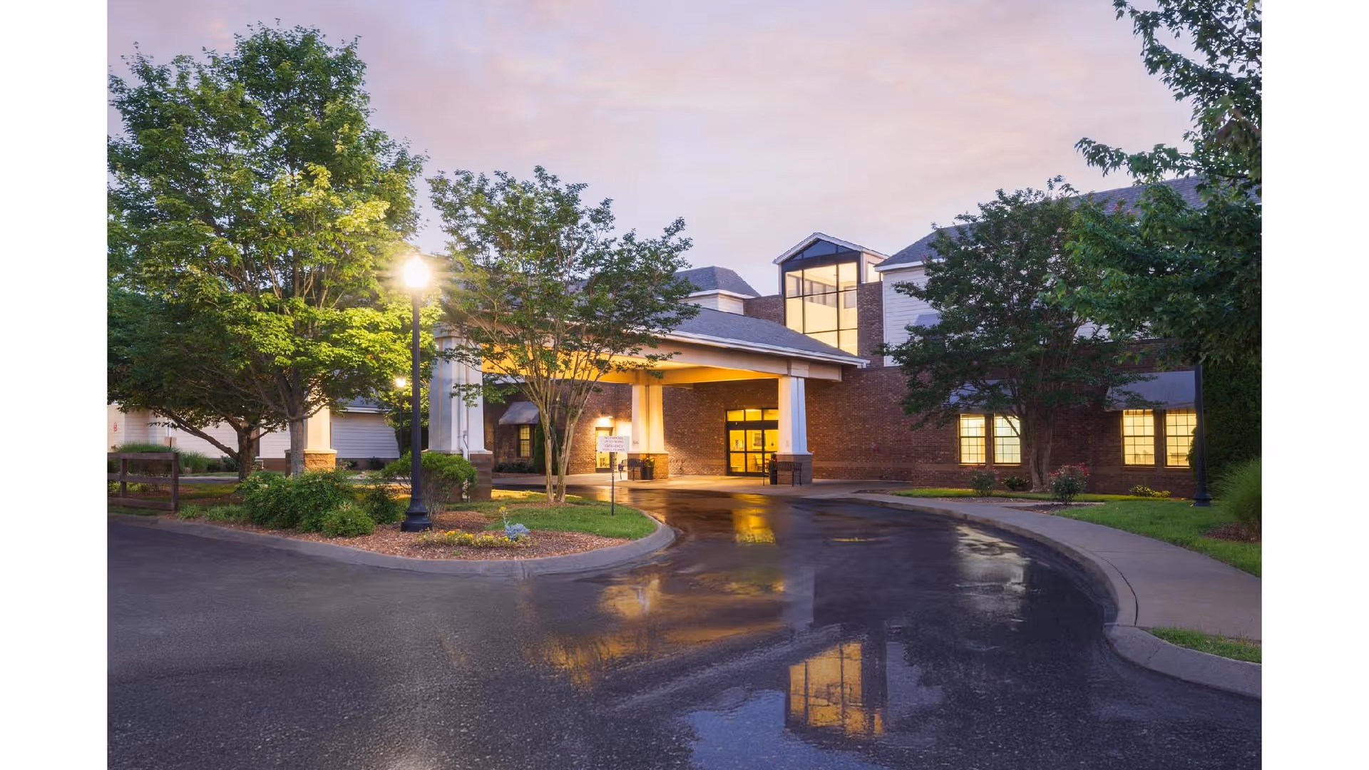 Covered main entrance of a senior living building at dusk with illuminated windows, trees, a lamppost, and a wet driveway reflecting lights.