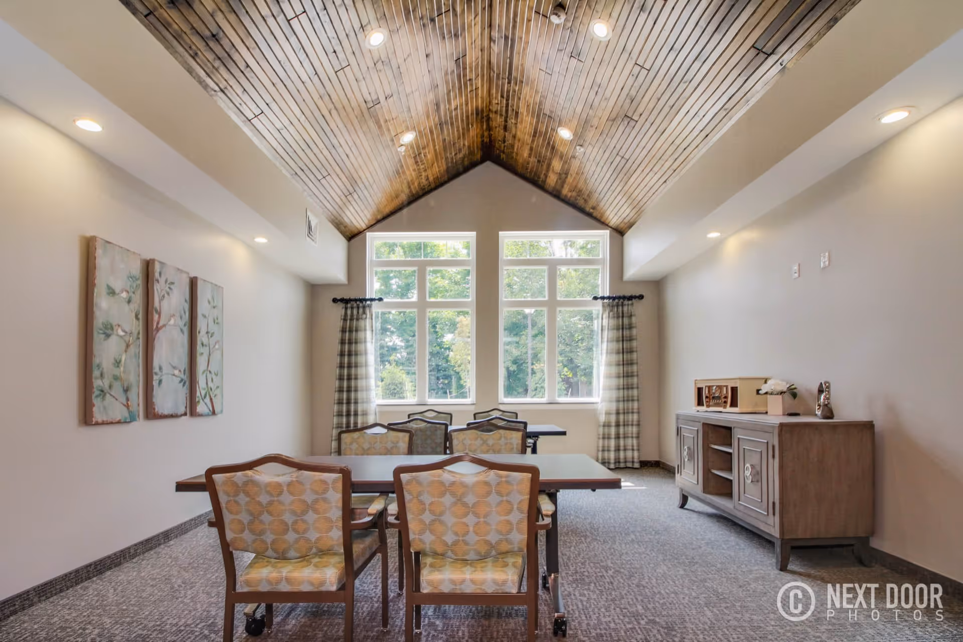 A bright room with a high wooden vaulted ceiling and large windows letting in natural light. The room contains a rectangular table with six patterned chairs around it, a wooden sideboard with decorative items on the right, and three framed artworks on the left wall. The windows have plaid curtains, and the carpet is gray.