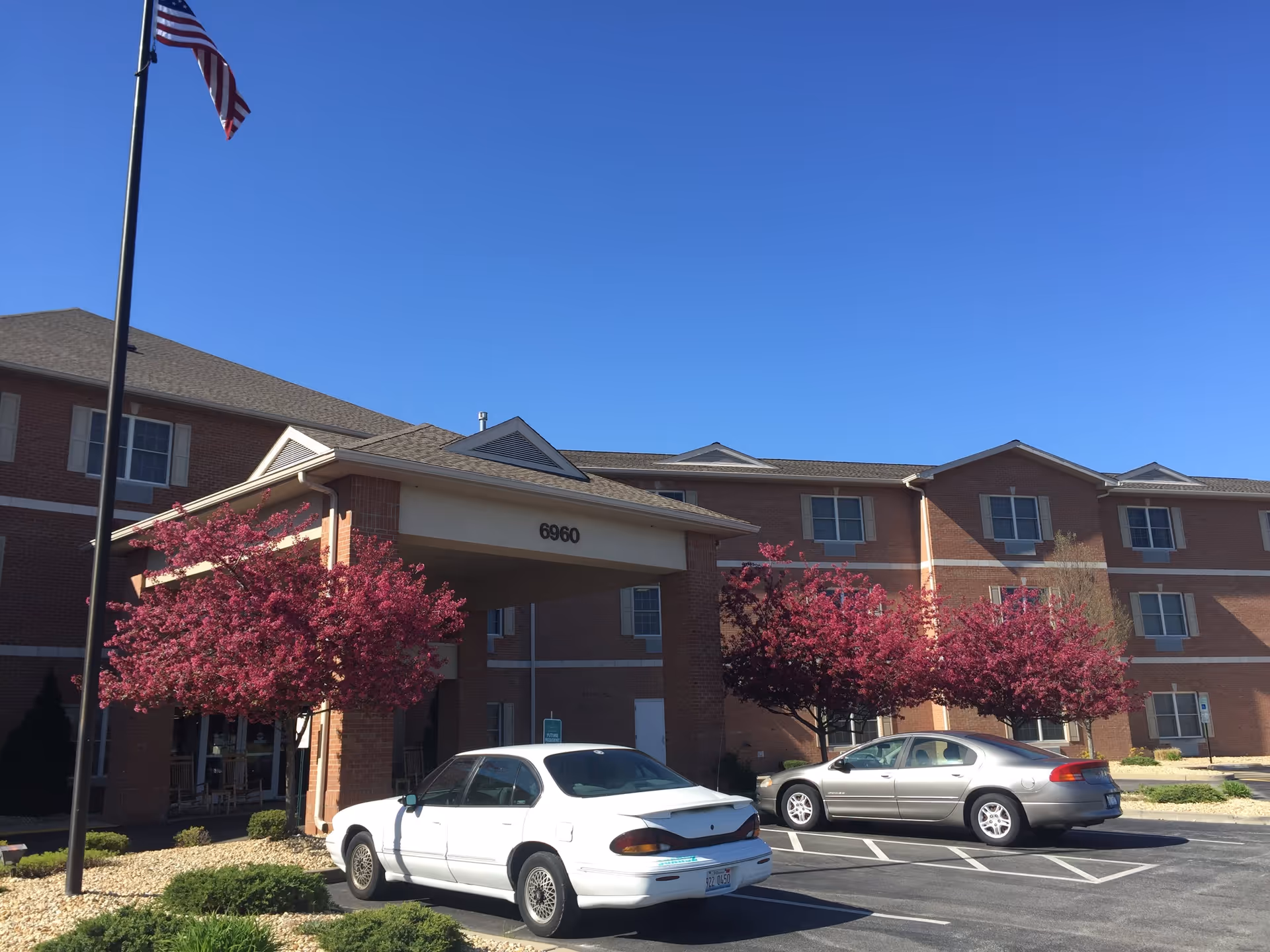 Exterior view of Cambridge House of Maryville, a multi-story brick building with several windows. There is a covered entrance with the number 6960 displayed. Two cars are parked in front, and there are blooming pink trees and a flagpole with an American flag near the entrance under a clear blue sky.