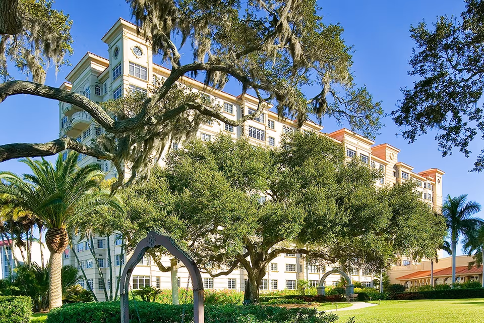 Exterior view of a multi-story senior living facility building with a clock tower, surrounded by large trees, palm trees, and well-maintained green lawns under a clear blue sky.