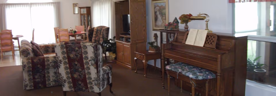 Communal living room with floral upholstered chairs, a piano and bench, TV cabinet, and a dining table visible in the background.