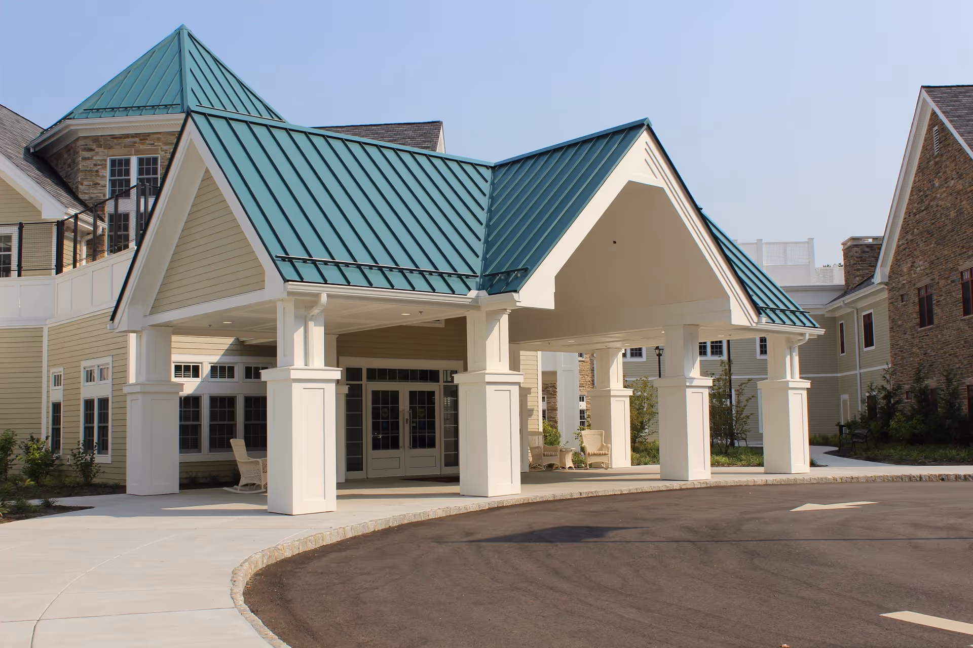 Entrance of a senior living facility with a covered drop-off area supported by white columns and a green metal roof. The building exterior features beige siding and stone accents, with windows and a paved driveway in front.