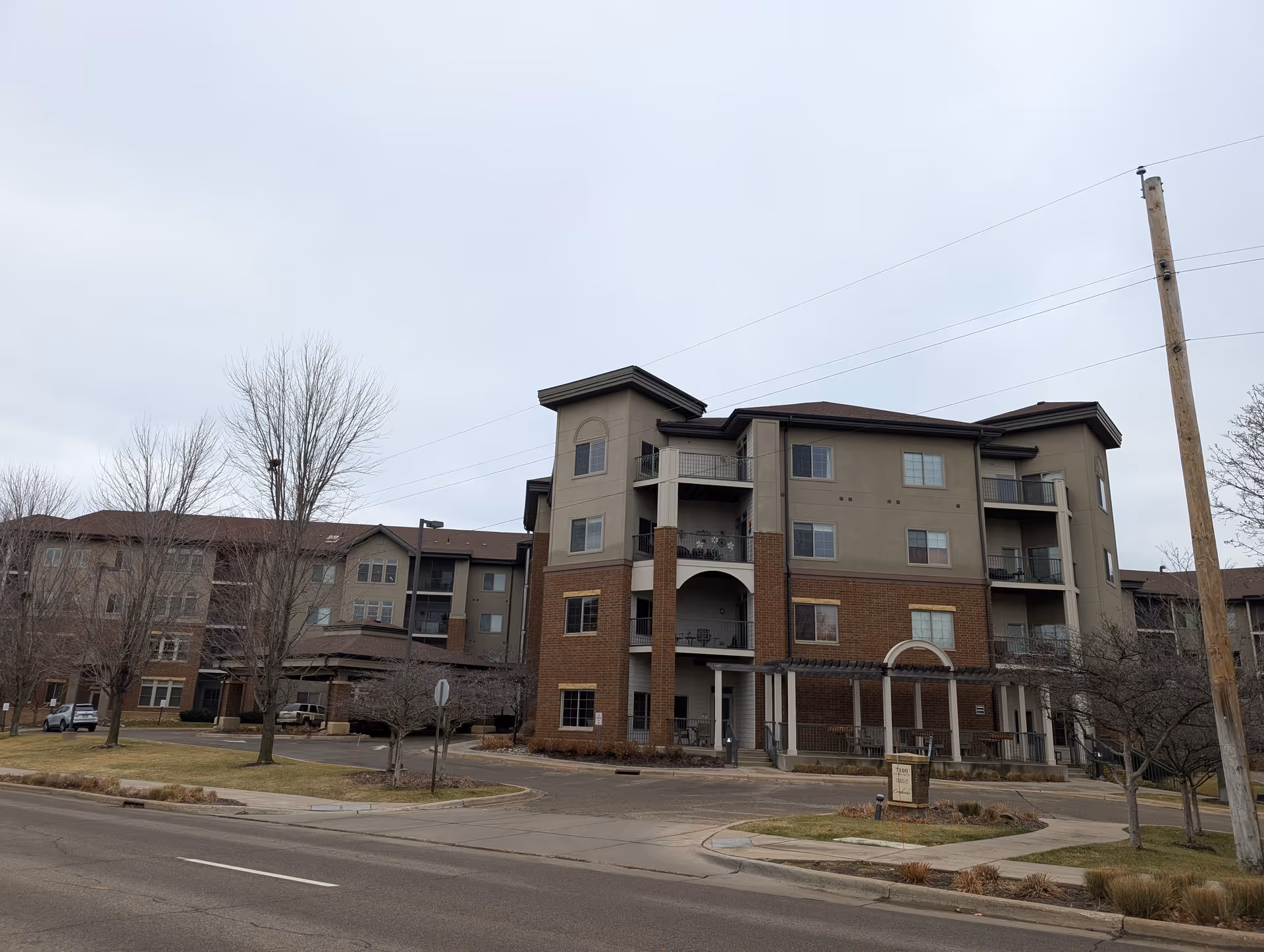 Exterior view of a multi-story senior living facility building with balconies, a covered entrance, and leafless trees in front. The sky is overcast and there is a street and sidewalk in the foreground.