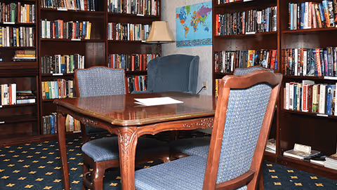 A cozy library room with wooden bookshelves filled with books lining the walls. In the center, there is a wooden table with four upholstered chairs around it. A lamp and a world map are visible on the wall behind the table.
