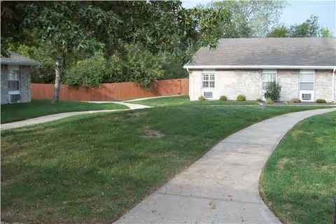 Curved walkway through green lawn leads to a brick senior living facility front entrance.