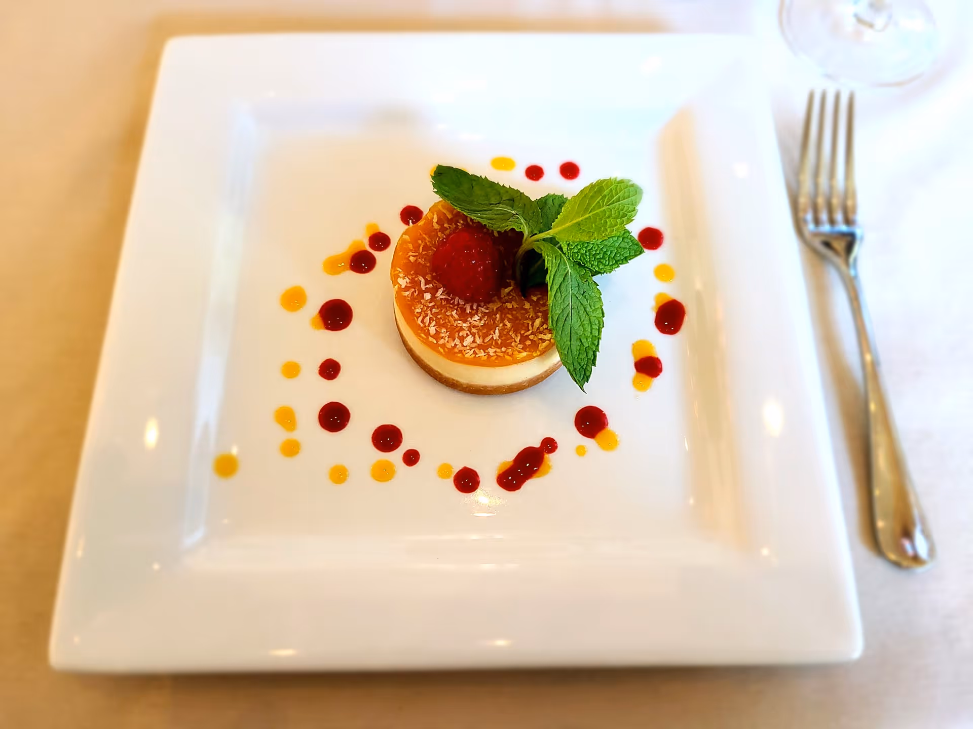 A small round cheesecake topped with a raspberry and mint on a square white plate decorated with red and yellow sauce dots and a fork at the side.