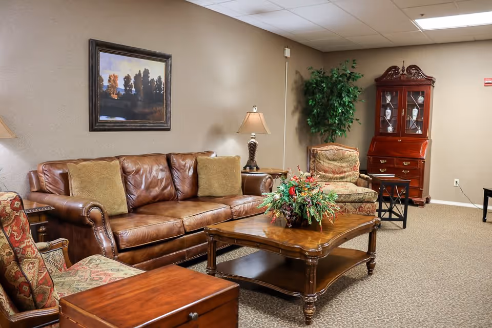 A cozy living room with a brown leather sofa, two patterned armchairs, a wooden coffee table with a floral arrangement, a side table with a lamp, a framed landscape painting on the wall, a tall green plant, and a wooden cabinet with glass doors displaying decorative items.