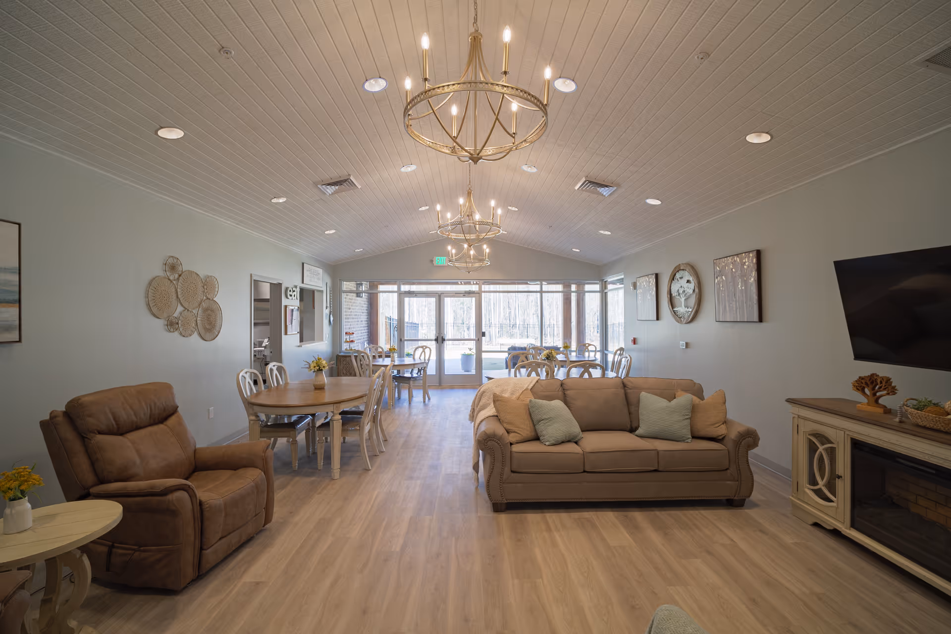 Spacious senior living common room with sofas, an armchair, dining tables, chandeliers, and large windows at the far end.