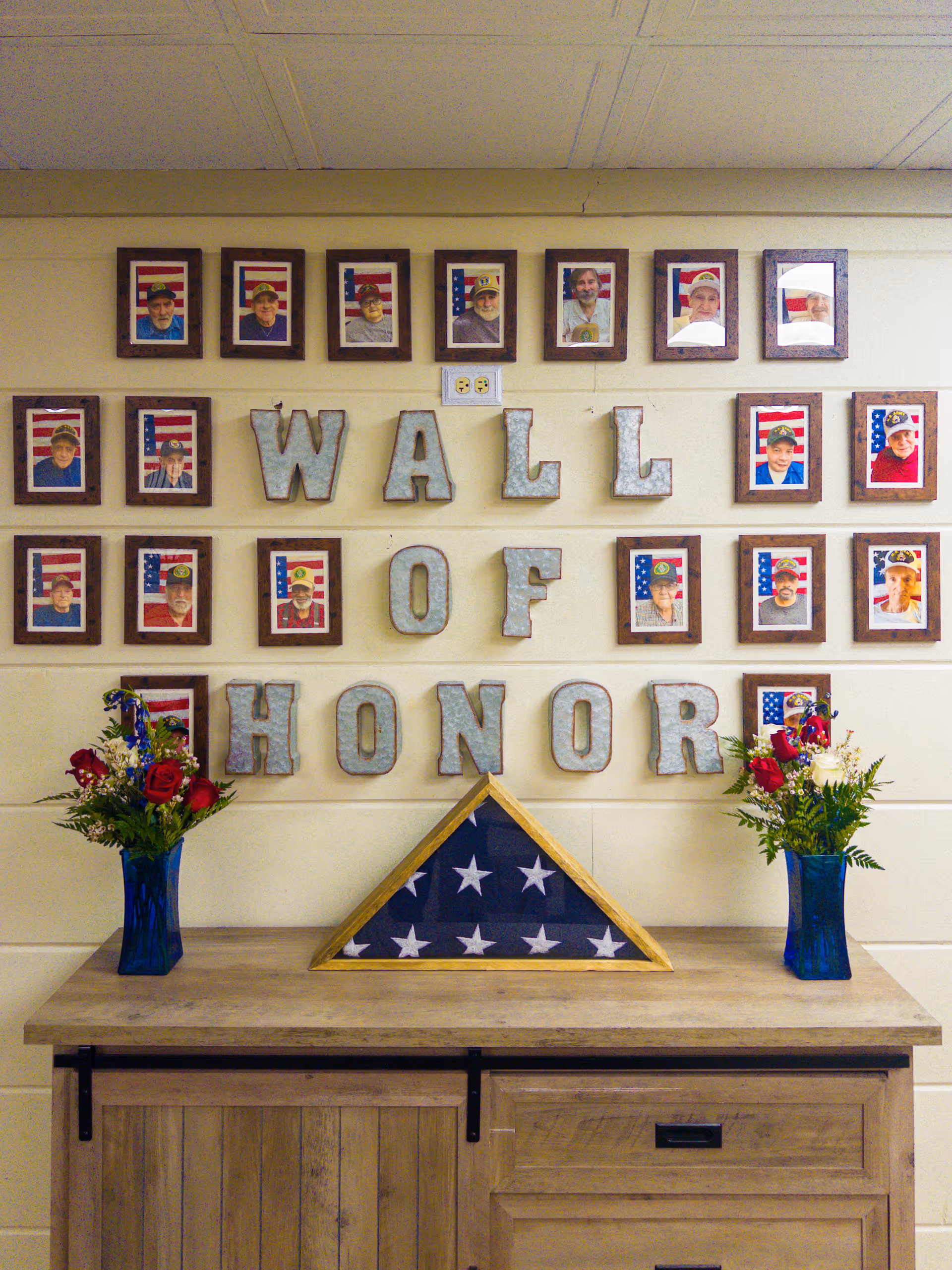 A wall display titled 'Wall of Honor' featuring framed photographs of veterans in front of American flags. Below the title are two vases with red, white, and blue floral arrangements and a folded American flag in a triangular wooden case placed on a wooden cabinet.