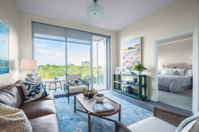 A bright living room with a brown leather sofa, two armchairs, a wooden coffee table, and a blue patterned rug. Large sliding glass doors open to a balcony with outdoor seating and a view of trees and buildings. A green console table with decorative items and a large colorful painting is against the wall. An open doorway leads to a bedroom with a neatly made bed.