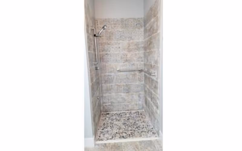 A tiled walk-in shower with a handheld showerhead mounted on a vertical bar and a horizontal grab bar on the right wall. The shower floor has small pebble-like tiles, while the walls are covered with larger rectangular tiles in neutral tones.